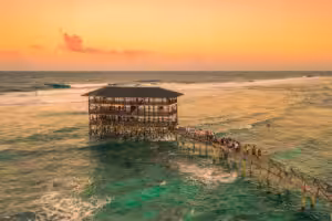 Overwater wooden café connected by a pier with people walking above turquoise ocean at sunset in the Philippines