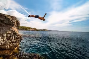 Man jumping from rocky cliff into clear blue ocean under sunny sky