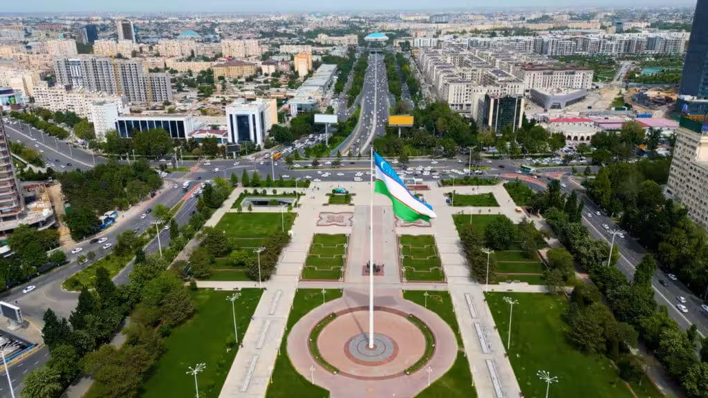 Aerial view of Independence Square in Tashkent with a large Uzbekistan flag flying on a tall flagpole, surrounded by formal gardens and wide tree-lined boulevards stretching across the city skyline