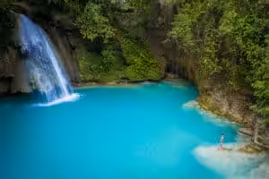 Kawasan Falls cascading into a vivid turquoise natural pool enclosed by moss-covered limestone cliffs and tropical jungle in Badian, Cebu, Philippines, with a lone visitor standing at the water's edge