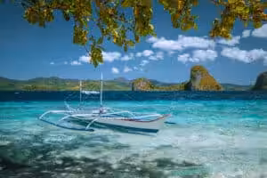 Traditional white Filipino bangka outrigger boat anchored in crystal-clear turquoise shallow water with dramatic limestone karst islands in the background, El Nido, Palawan, Philippines