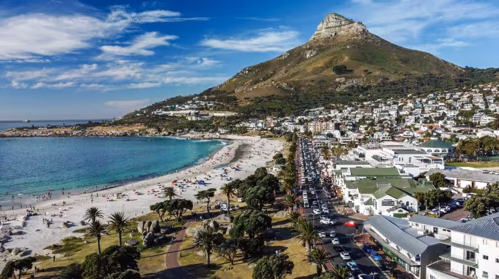 Aerial view of Camps Bay beach with white sand, turquoise water, palm-lined promenade and Lion's Head mountain rising behind the upscale beachfront suburb of Cape Town, South Africa