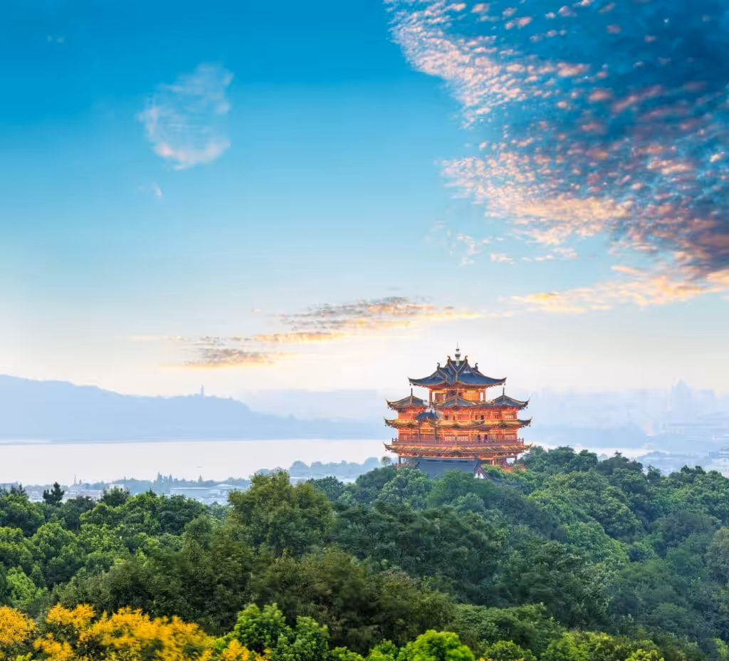 The illuminated Chenghuang Pavilion rising above a dense green forest with West Lake and the Hangzhou city skyline visible in the misty background at sunrise, Zhejiang Province, China