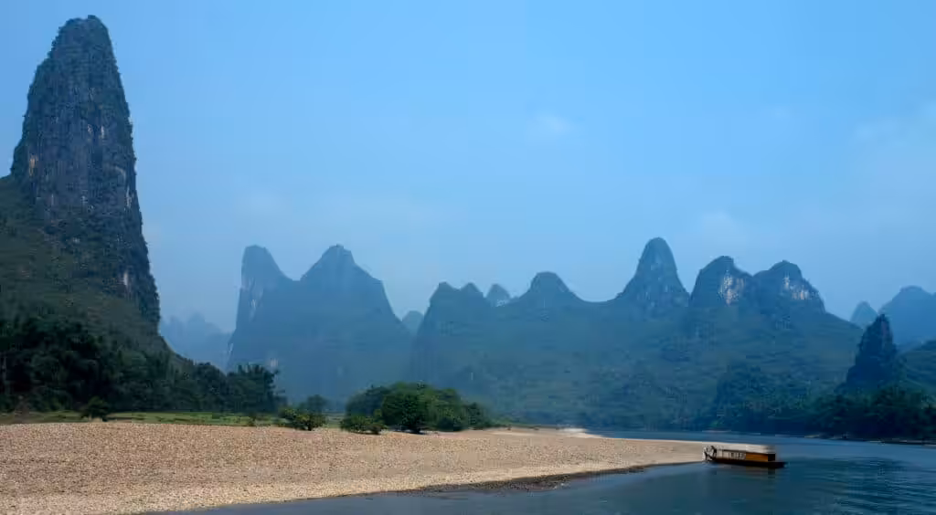 A traditional wooden river boat cruising along the Li River with dramatic karst limestone peaks rising from the misty green valley near Guilin, Guangxi, China