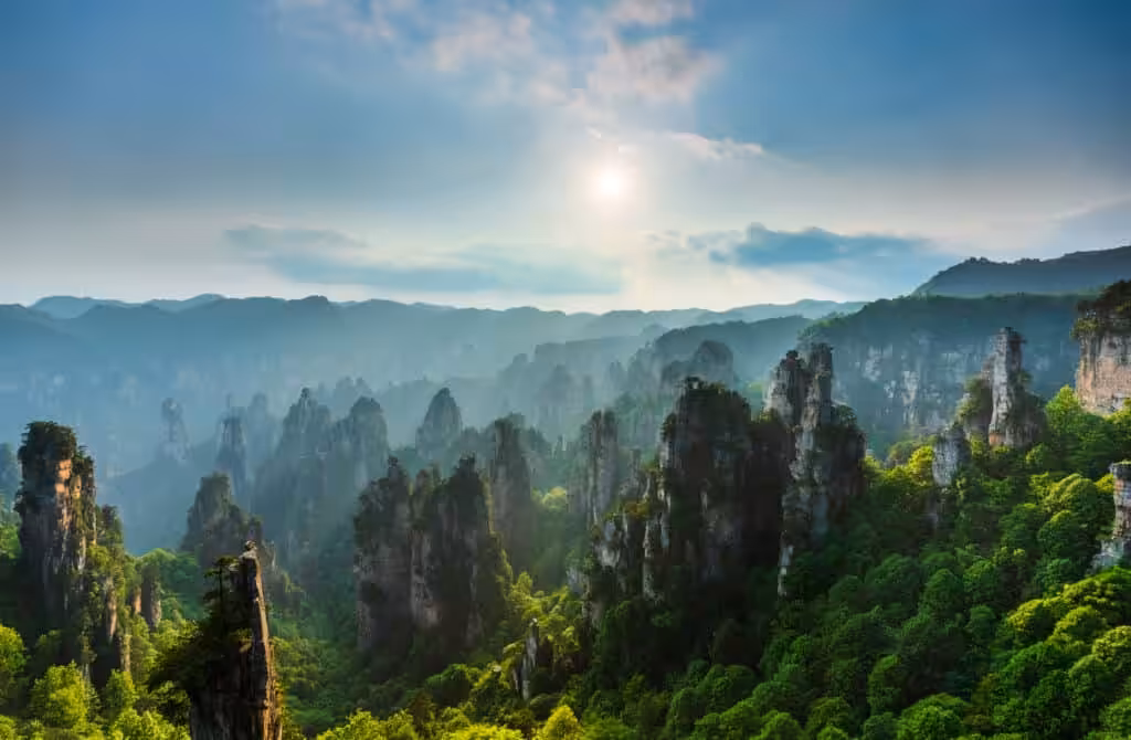Panoramic sunset view over hundreds of towering sandstone pillar formations rising from a lush green forest valley with sunlight breaking through clouds and mountain ranges fading into the haze, Zhangjiajie National Forest Park, Hunan, China